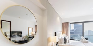 Habitación de hotel moderna con un espejo grande y redondo, cama con ropa blanca, televisor de pantalla plana y vistas a la ciudad a través de ventanas amplias.