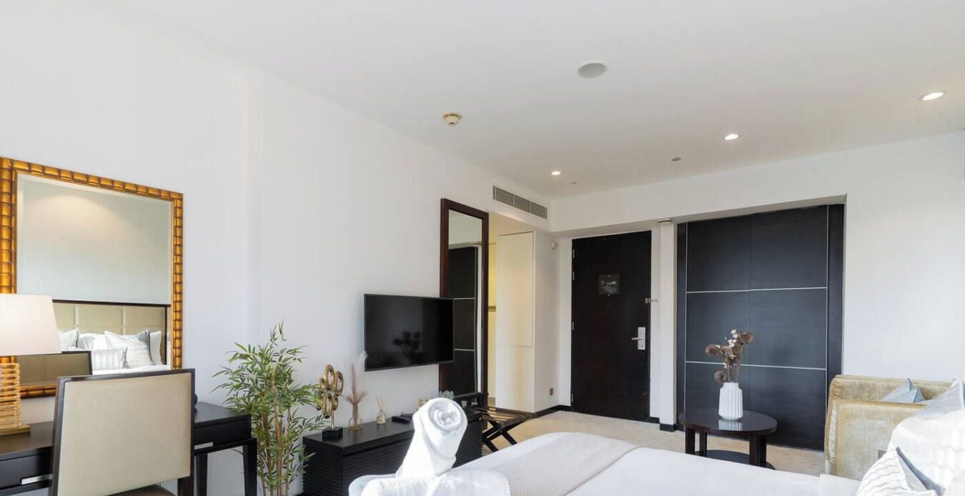 A hotel room with a bed, a desk, a chair, a TV, a mirror, and a door leading to another room.
