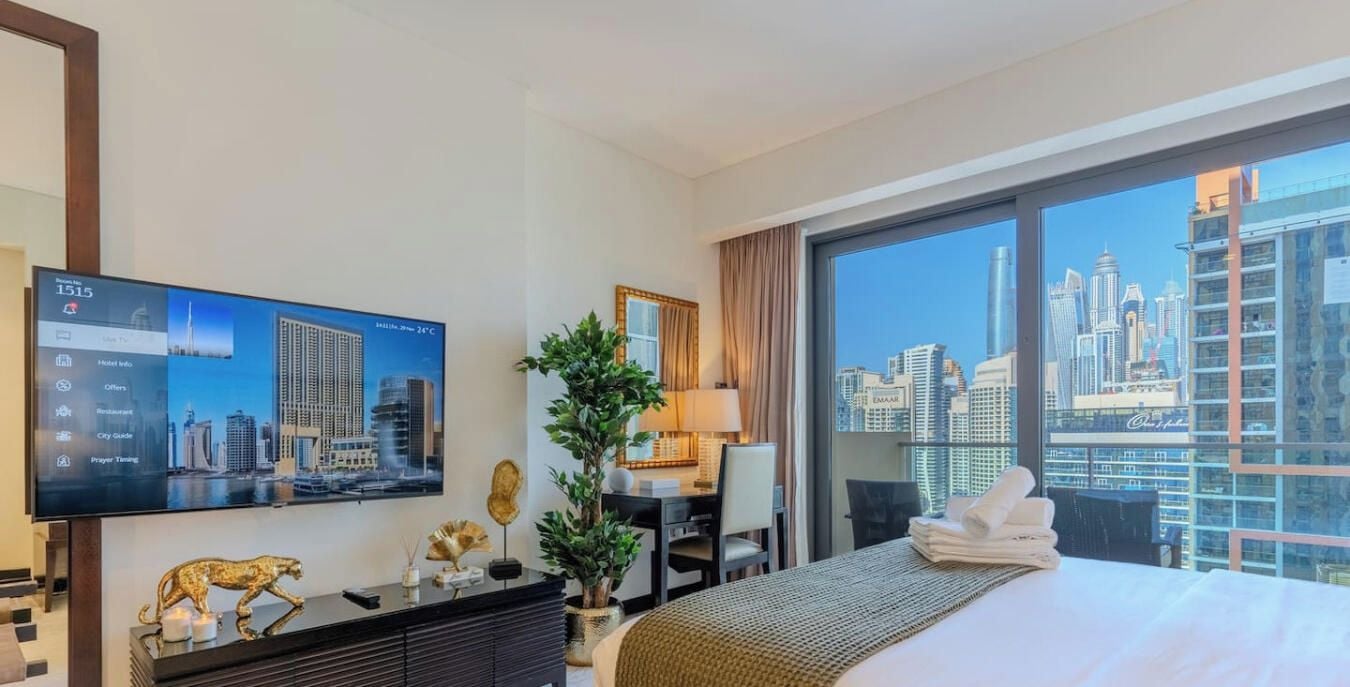 Habitación de hotel con un ventanal grande, cama, escritorio, televisor y vista a la ciudad.