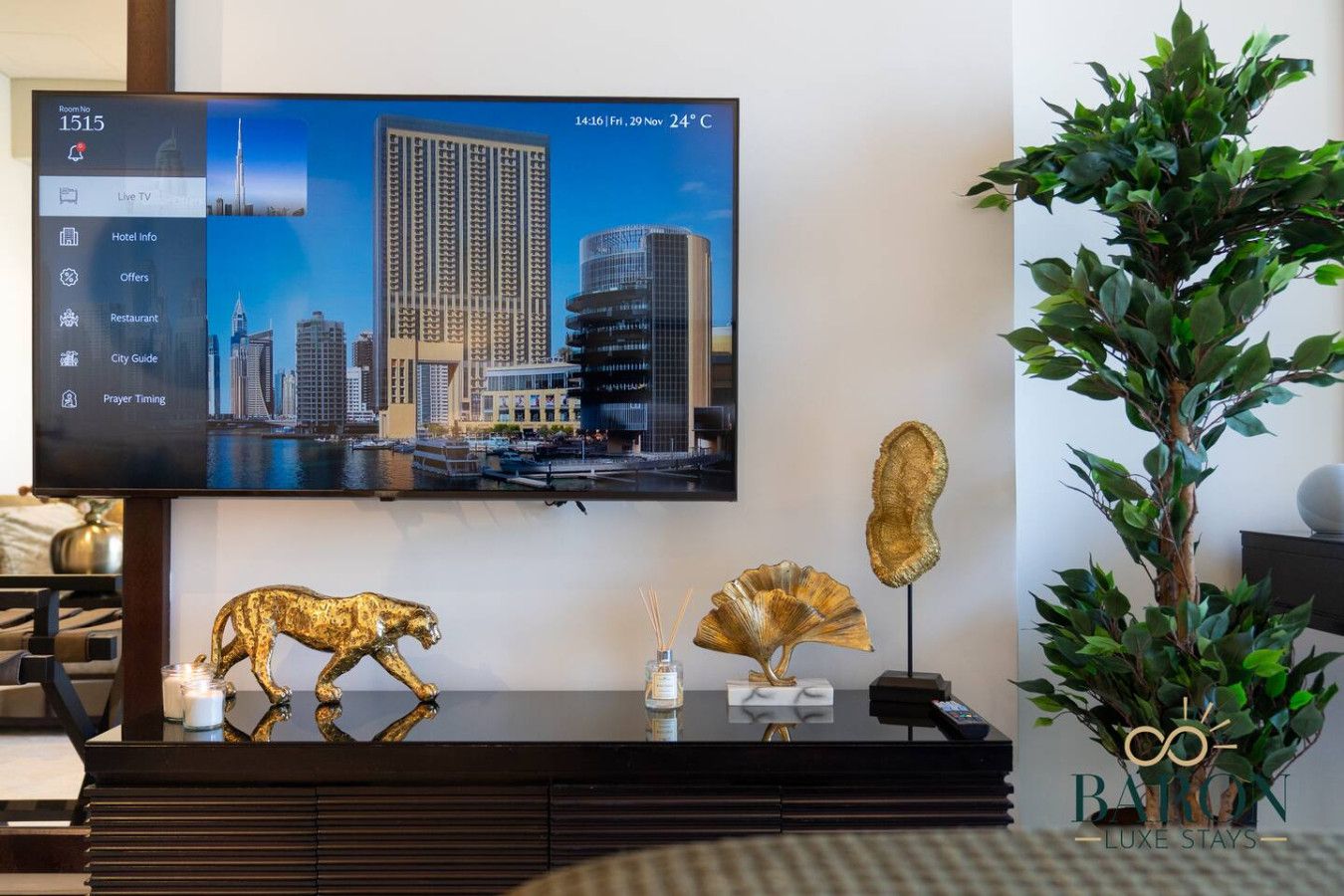 Un televisor mostrando un paisaje urbano con una pantalla de información de hotel, una figura dorada decorativa de un animal y una planta en maceta.