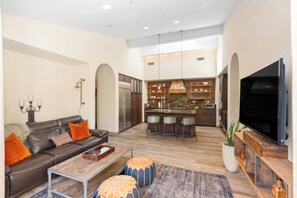 A spacious living room with a leather sofa, ottomans, and a coffee table. A kitchen with a large island and bar stools is visible in the background. - 3-bedroom estate in SantaLuz Community  with AC and fitness room and Spa (San Diego)