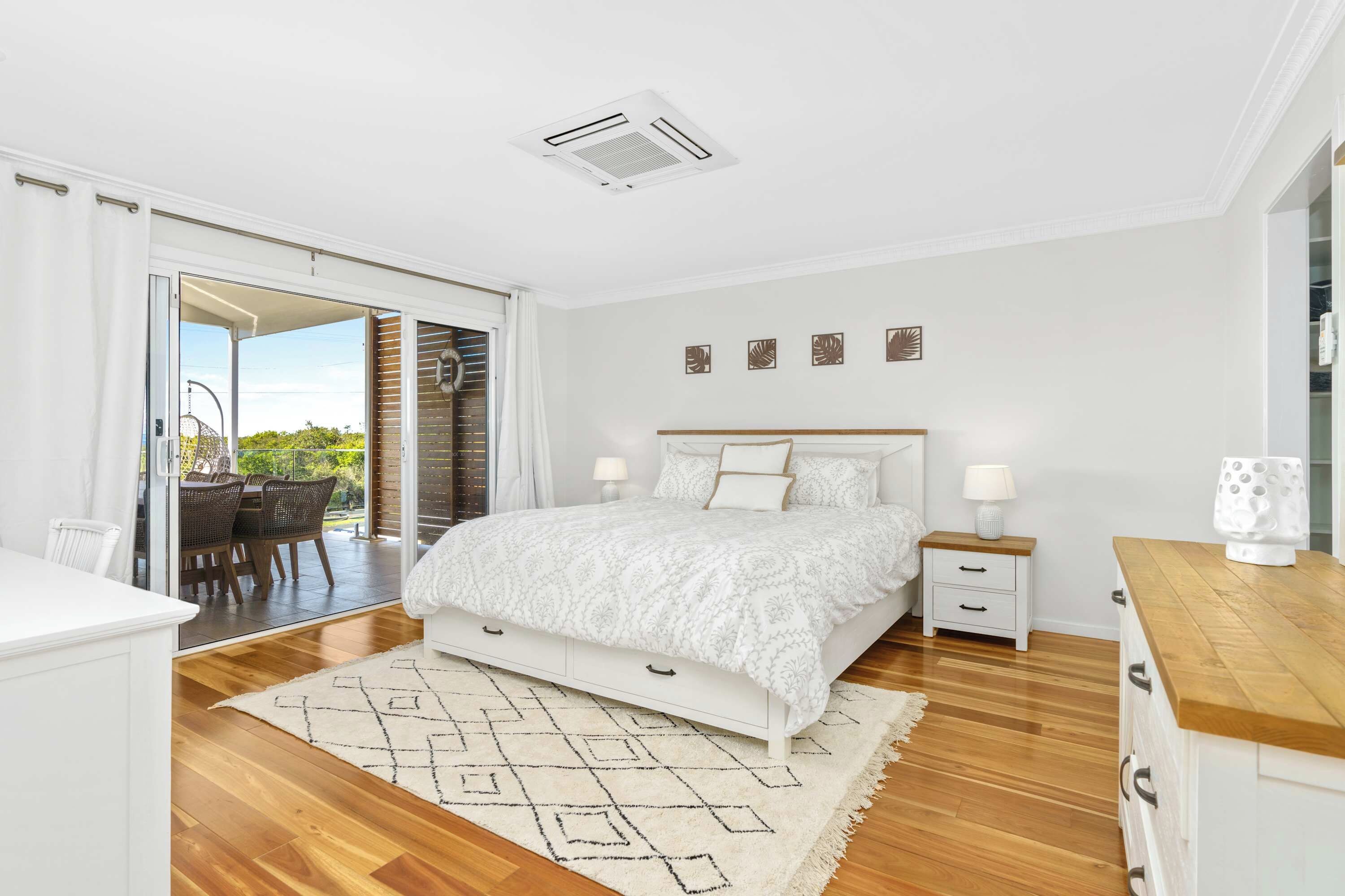 A bedroom with a bed, bedside tables, a nightstand, a wooden floor, a patterned rug, and a view through a sliding door to an outdoor area.