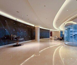 A modern lobby with a curved glass wall, a marble wall with a decorative table, and a seating area with sofas. - The Exquisite • JW Marriott (Dubai)