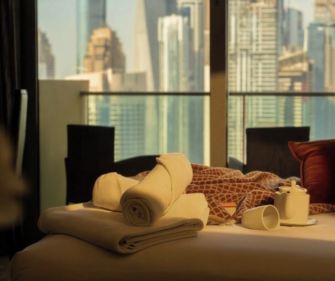 A hotel room with a bed, pillows, a rolled towel, and a cup on the bedside table.