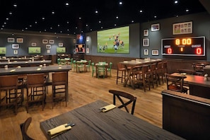 A sports bar with multiple seating areas, a large screen displaying a sports event, and a digital scoreboard. - The Urban • JW Marriott (Dubai)