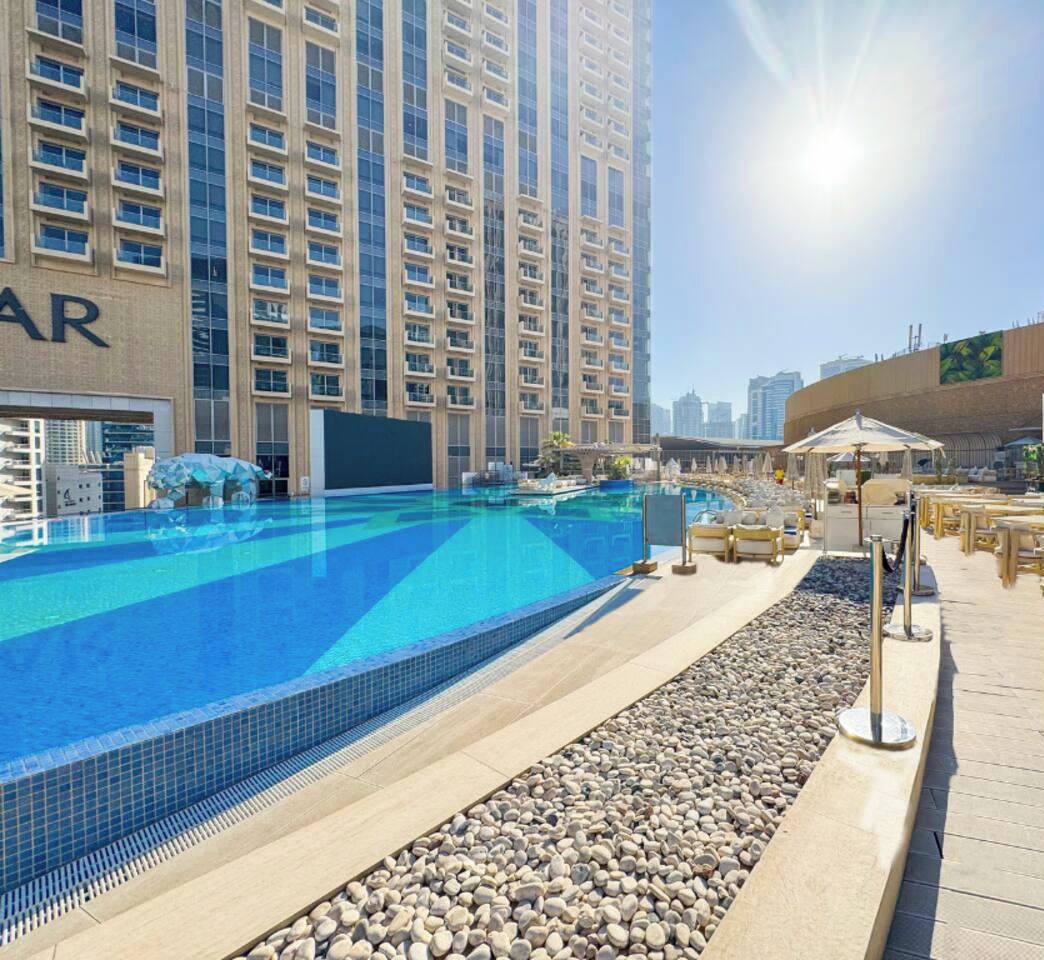 Ein Schwimmbeckenbereich mit blauem Himmel, ein Gebäude mit einem sichtbaren 'AR'-Schild und ein sonniger Sitzbereich im Freien.