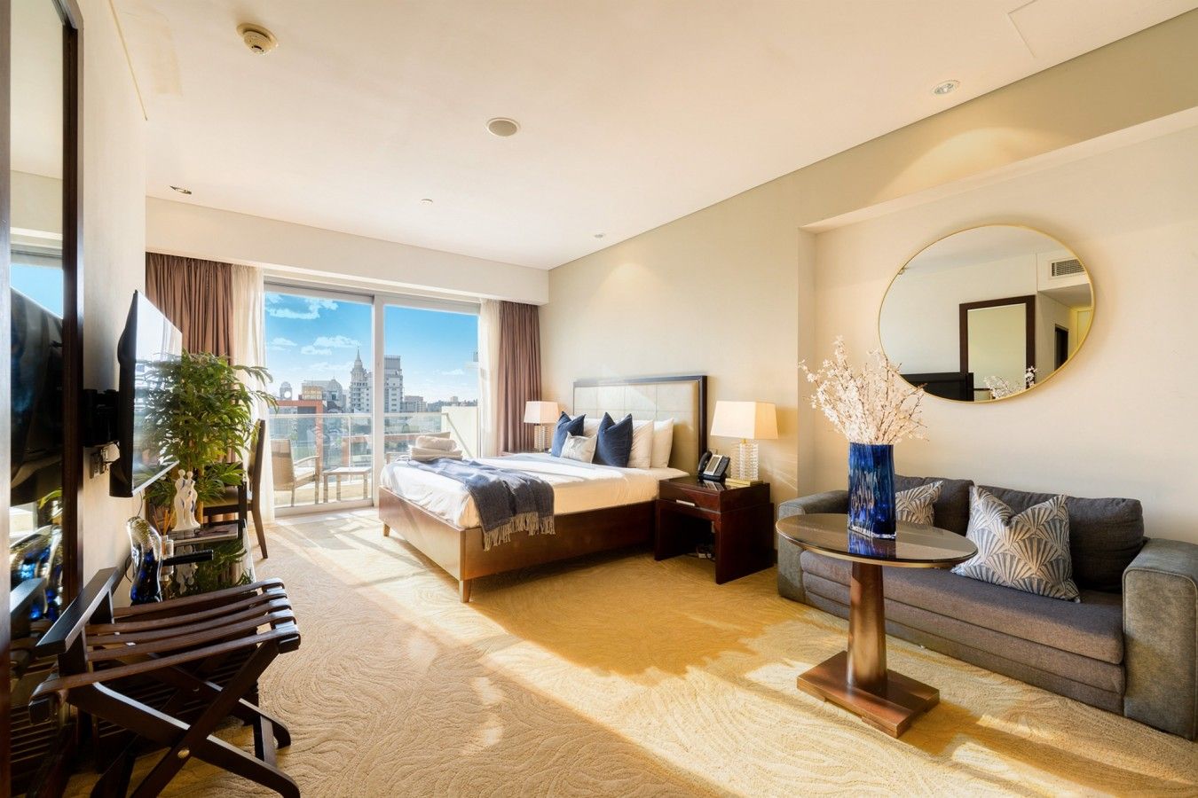 A hotel room with a bed, a sofa, a TV, a desk, and a view of the city.