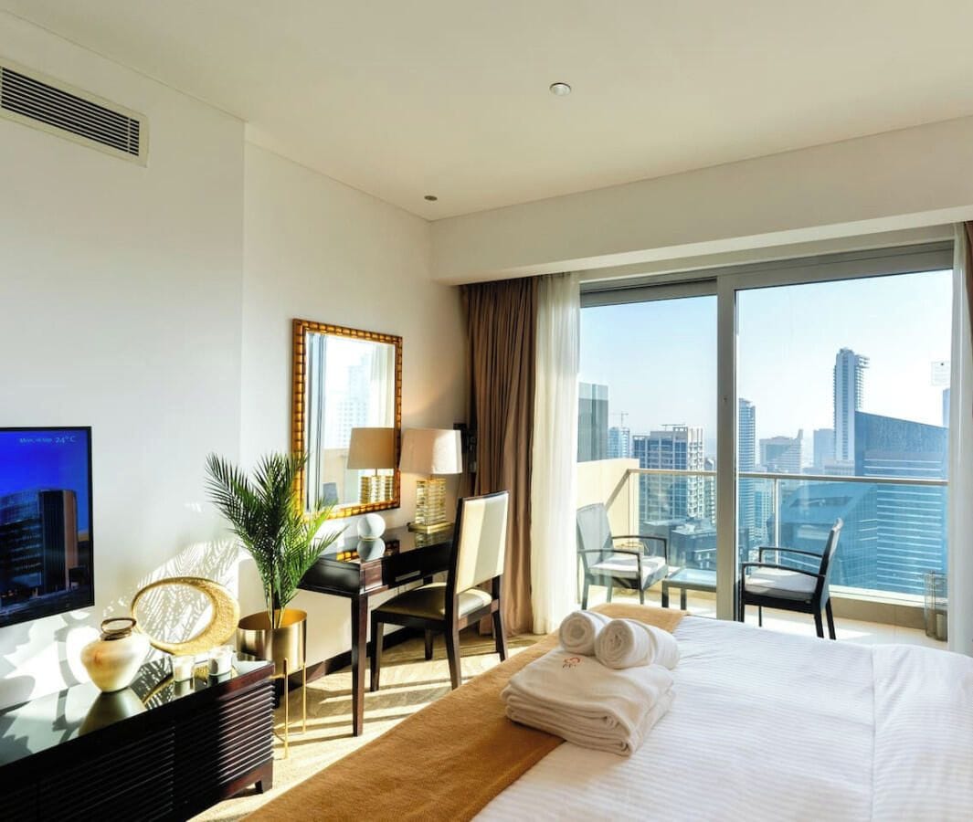 Une chambre d’hôtel équipée d’un lit, d’un bureau, d’une chaise, d’une télévision et d’une grande fenêtre donnant sur la ville.
