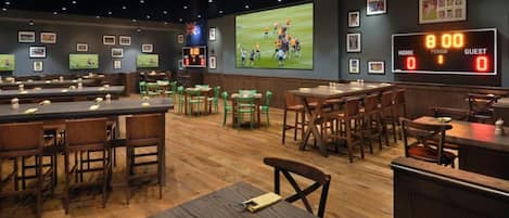 Un bar sportif doté de plusieurs espaces pour s’asseoir, d’un grand écran diffusant un événement sportif et d’un tableau de bord numérique.