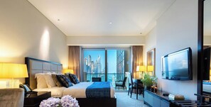 Ein Hotelzimmer mit Bett, Nachttisch, Lampe, Fernseher und Blick auf eine Stadtlandschaft durch große Fenster.