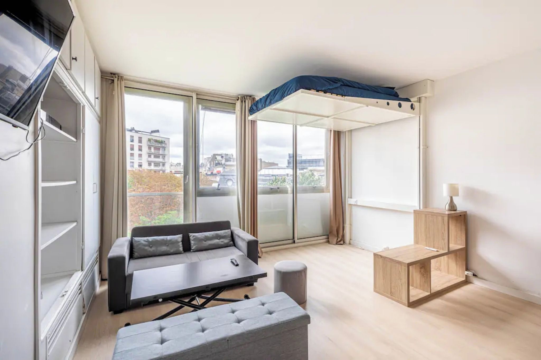 Un appartement moderne doté d’un canapé, d’un lit convertible, d’une petite table d’appoint en bois et offrant une vue sur la ville grâce à de grandes fenêtres.