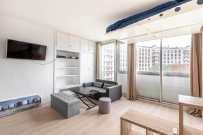A modern living room with a sofa, ottoman, and a flat-screen TV mounted on the wall. - Bright Studio-15 Minutes from the Arc de Triomphe (Paris)