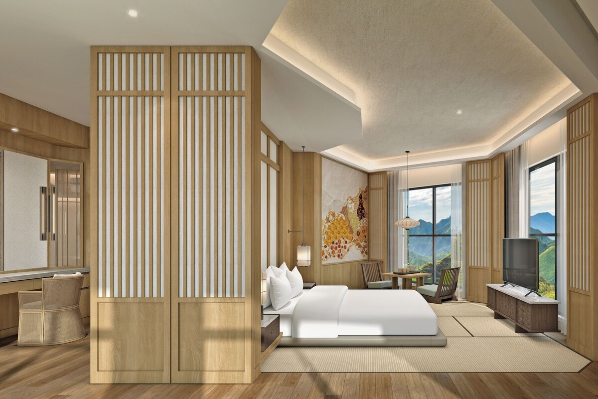 A modern hotel room with a large bed, a desk, a TV, and a view of mountains through the window.