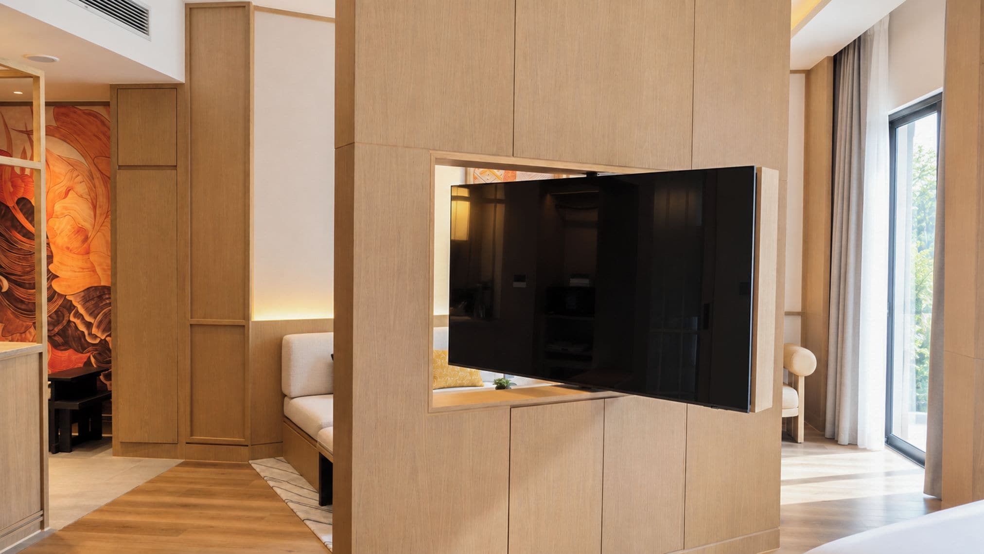 Habitación de hotel moderna con televisión de pantalla plana montada en un panel de madera, un sofá y un ventanal con cortinas.