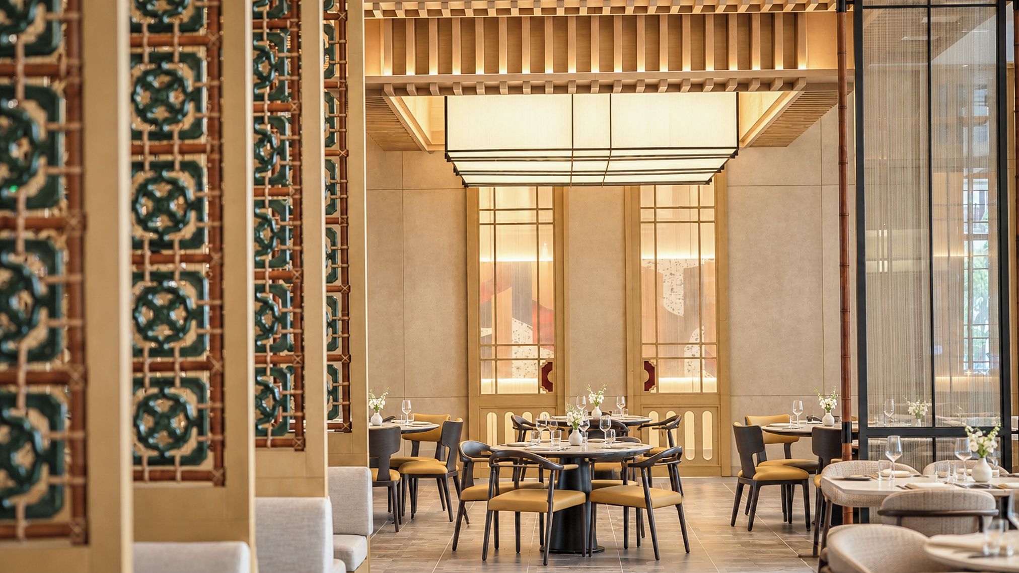 A restaurant interior with multiple dining tables and chairs, a decorative wall with a geometric pattern, and a large illuminated ceiling feature.
