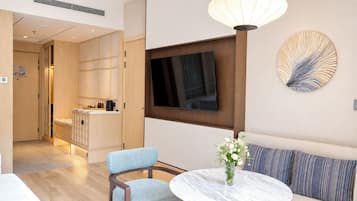 A modern hotel room with a dining area, a sofa, a TV mounted on the wall, and a decorative wall piece.