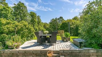 An outdoor patio with a table and chairs, surrounded by greenery and a clear sky.