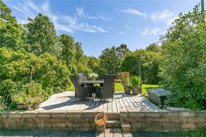 An outdoor patio with a table and chairs, surrounded by greenery and a clear sky.