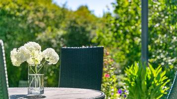 Mesa redonda con un florero de vidrio con flores blancas, una silla verde oscuro y un jardín con diversas plantas al fondo.