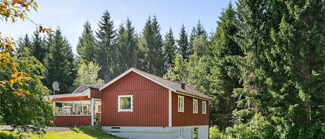Una casa roja de dos pisos con techo inclinado, rodeada por un césped verde exuberante y un bosque denso.