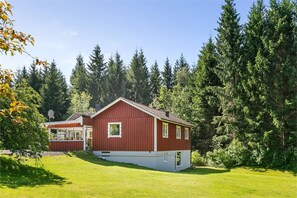 A red two-story house with a sloped roof, surrounded by a lush green lawn and dense forest.