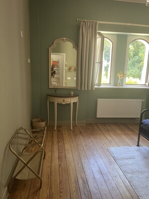 Una habitación con suelo de madera, un espejo, una mesita, un radiador y una ventana con cortinas.