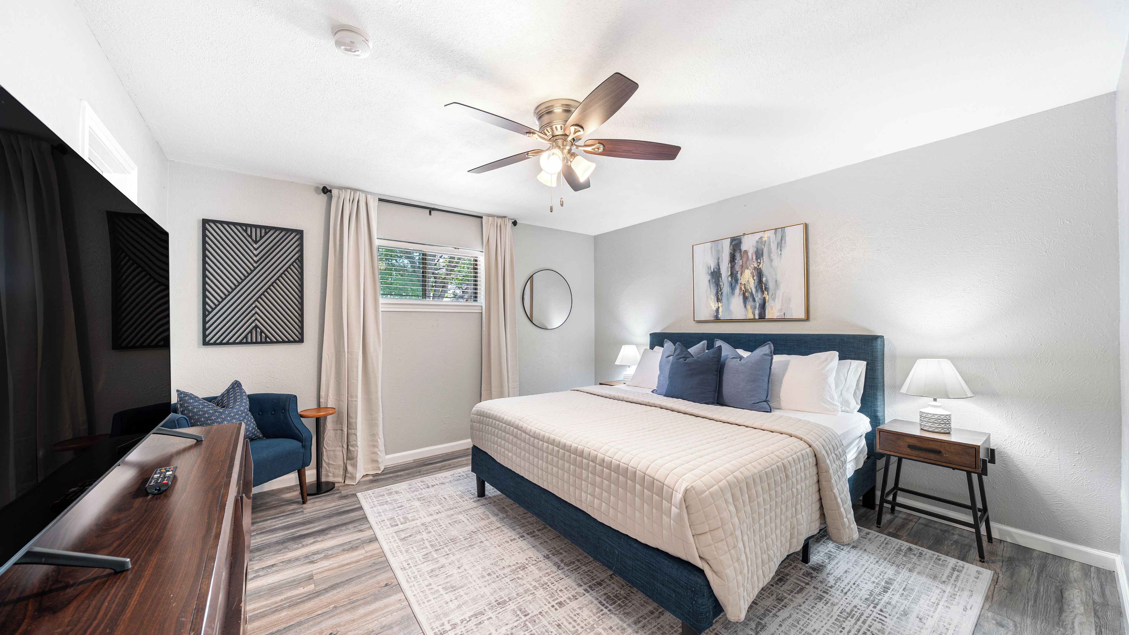 A hotel room with a bed, a desk, a chair, a ceiling fan, a window with curtains, and artwork on the walls.