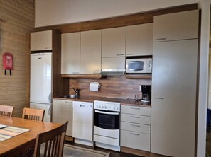 A compact kitchen with a stove, microwave, and refrigerator, wooden cabinets, and a dining table with chairs. - Länsirinne F 24 | 2BR Apt in Ruka Centre (Kuusamo)