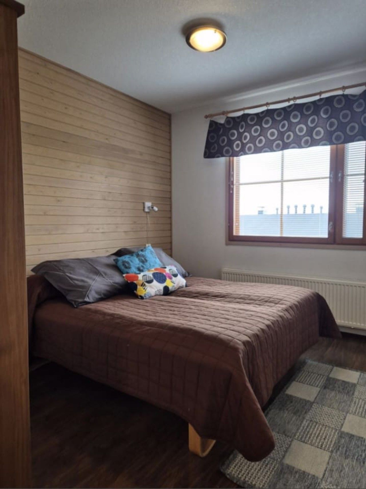 Una camera da letto con una testiera in legno, un letto con copripiumino marrone, una finestra con vista e una tenda a motivi.