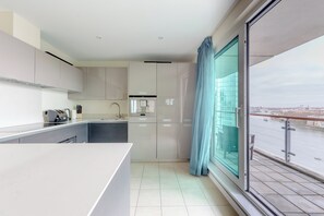 A modern kitchen with a large island, a dining area, and a view of a river and cityscape. - Sleek 3BD flat with Stunning City Views, Vauxhall (London)