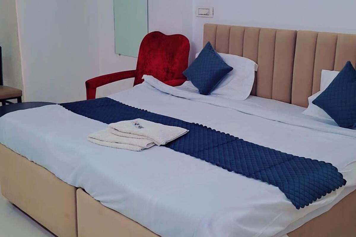 Uma cama bem arrumada com cabeceira, uma cadeira vermelha e uma parede branca com interruptor de luz.