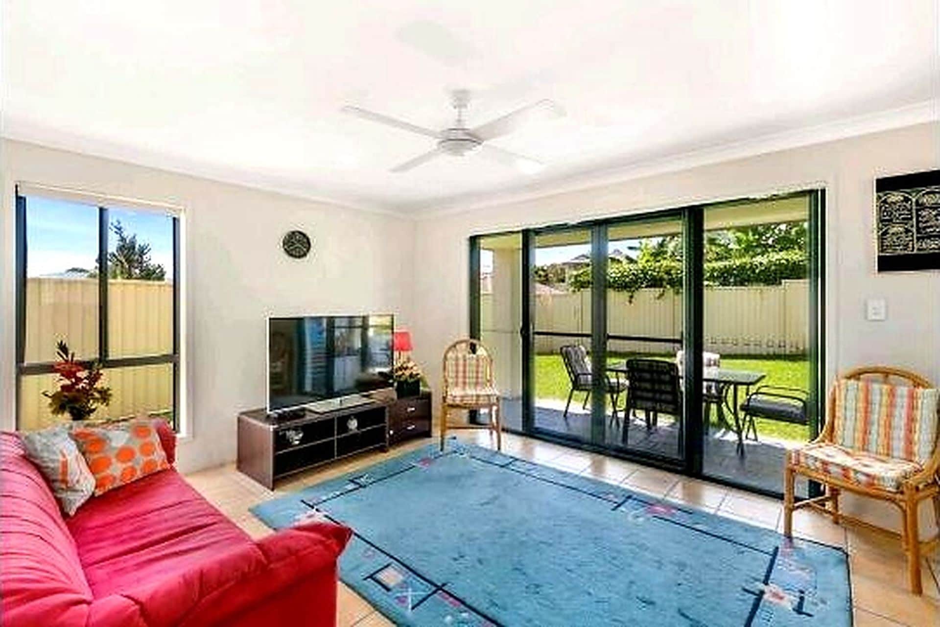 A living room with a red sofa, a TV, and a coffee table. There is a sliding glass door leading to an outdoor patio with chairs and a table.