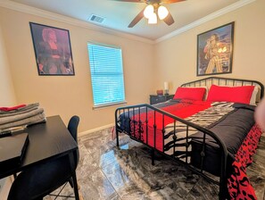 A bedroom with a bed, a desk, a chair, a window with blinds, a ceiling fan, and two framed pictures on the wall. - Private World for two, one bedroom house with music & arcade games. (Houston)