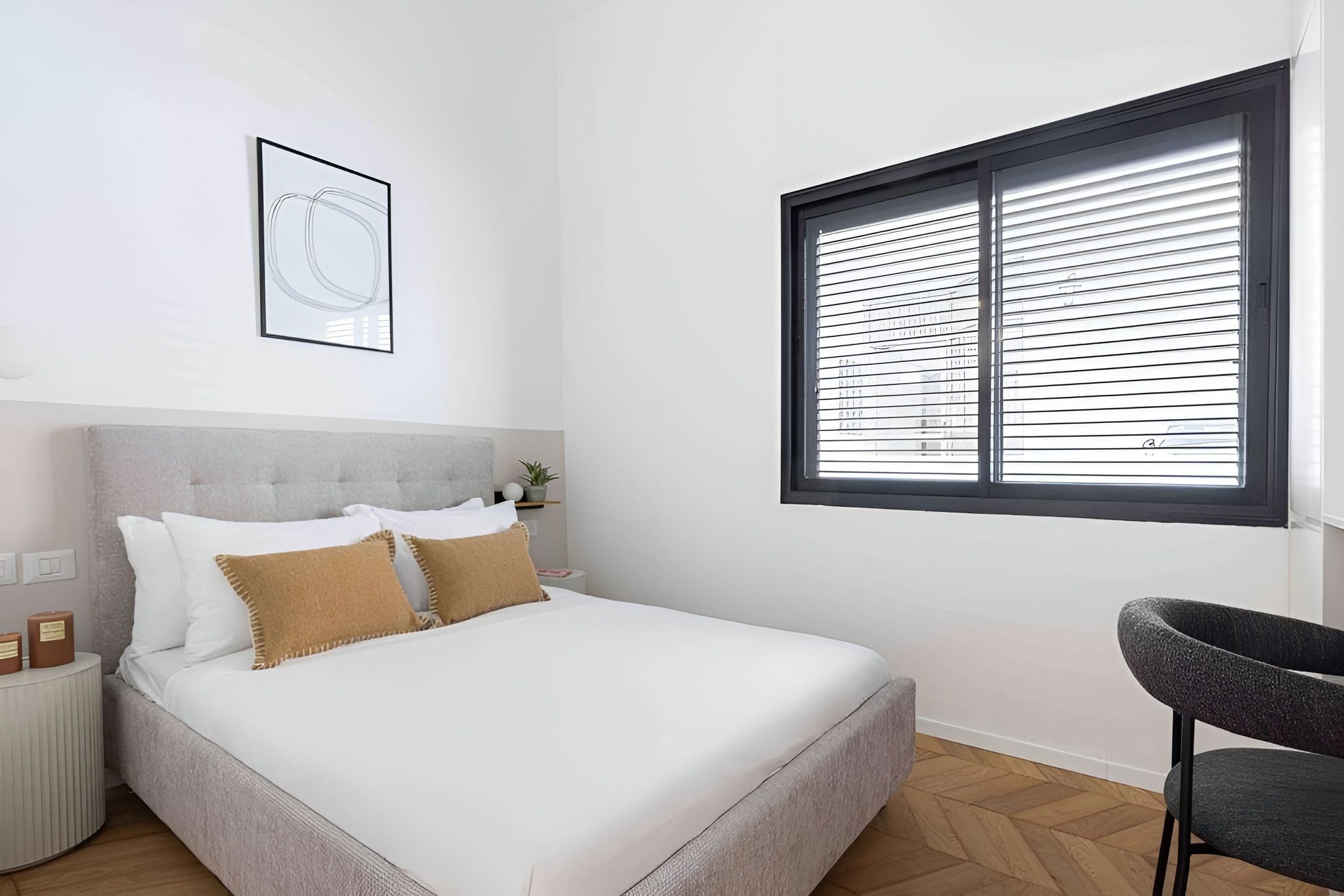 Un dormitorio con cama, una silla, una ventana con persianas, un cuadro en la pared y una mesita con una planta.