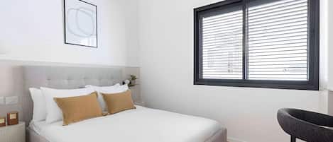Un dormitorio con cama, una silla, una ventana con persianas, un cuadro en la pared y una mesita con una planta.