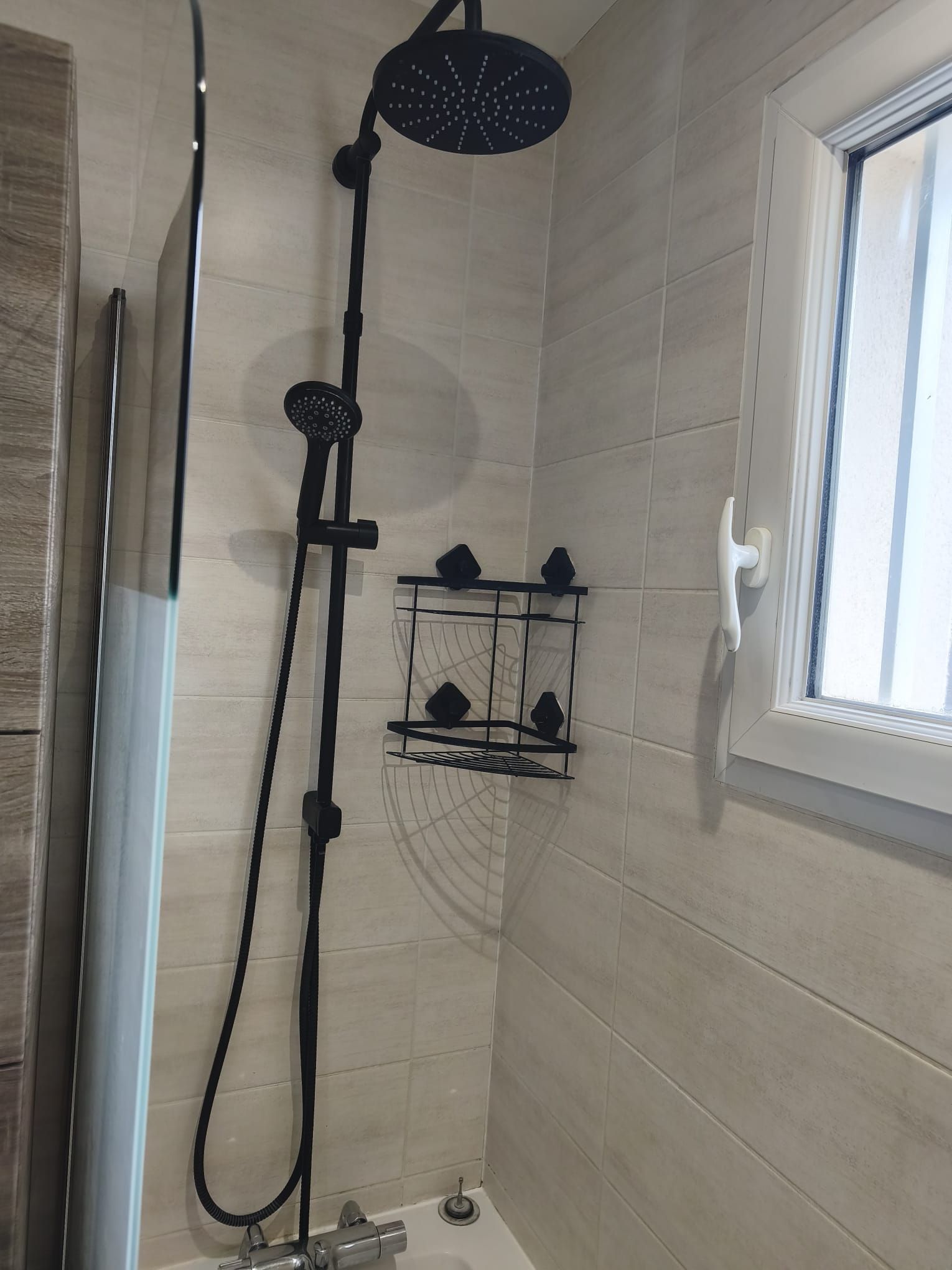 Un espace douche équipé d’un pommeau de douche à main, d’une étagère fixée au mur pour les articles de toilette et d’une fenêtre à cadre blanc.