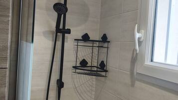 Un espace douche équipé d’un pommeau de douche à main, d’une étagère fixée au mur pour les articles de toilette et d’une fenêtre à cadre blanc.