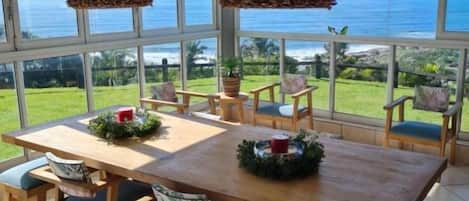 A dining area with a large table and chairs, large windows with a view of the ocean, and hanging light fixtures.