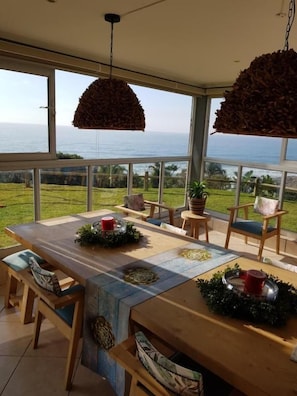 An outdoor dining area with a view of the ocean, a wooden table, chairs, and hanging light fixtures. - Alomsee 20 Ramsgate 4 Bedroom Apartment (Margate)