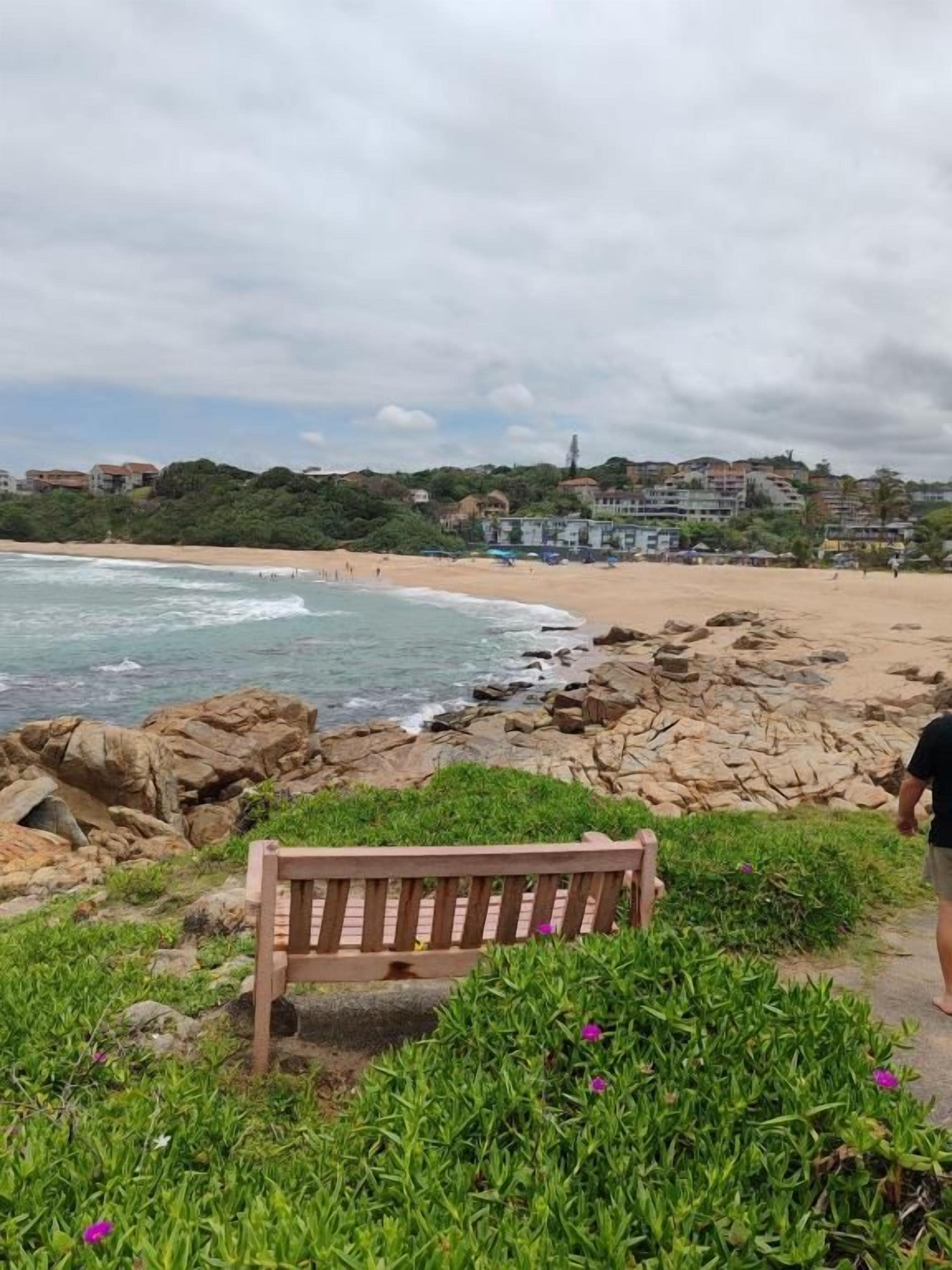 A beach with a wooden bench, rocky shoreline, and buildings in the background.