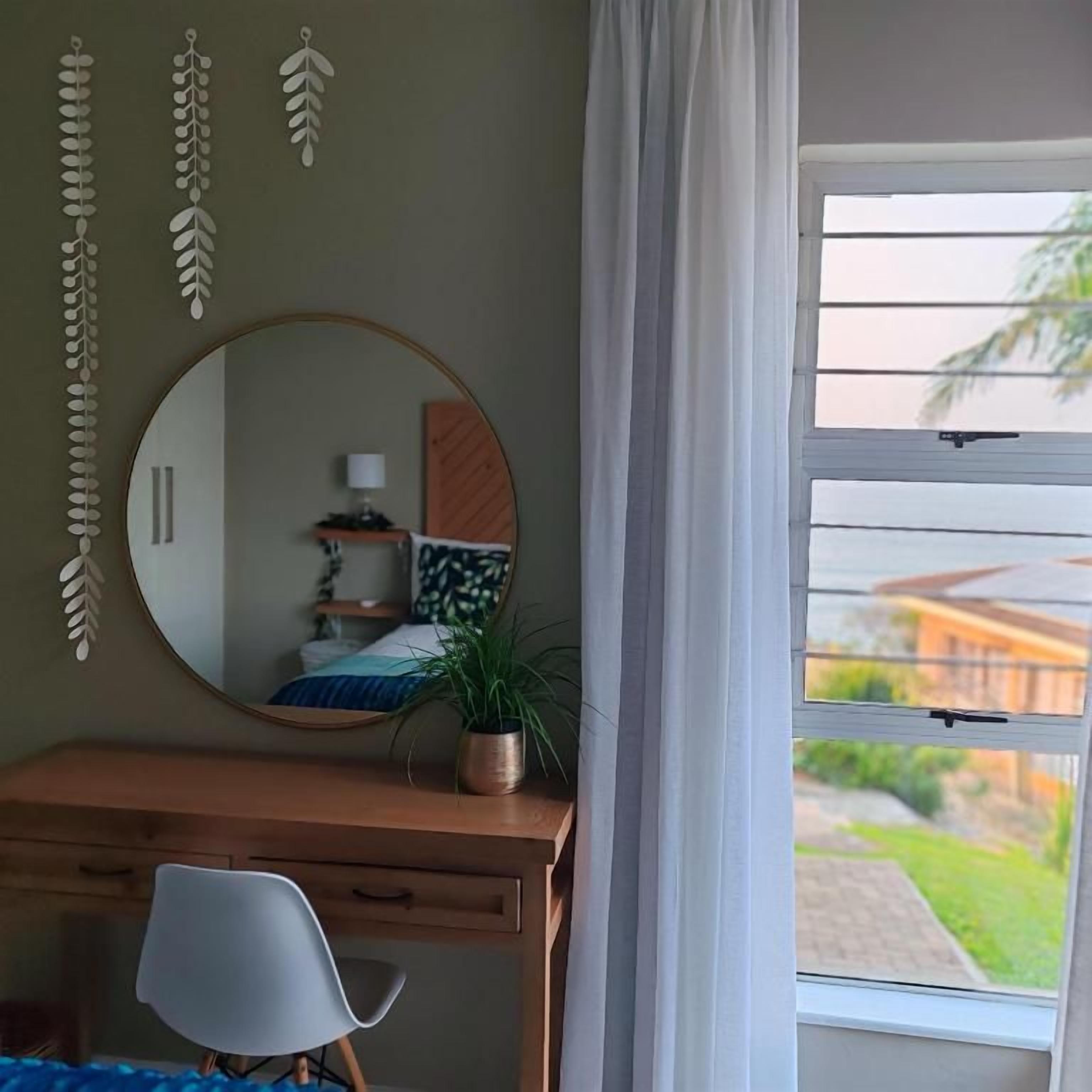 A bedroom with a wooden desk, a chair, a round mirror, a potted plant, and a window with blinds.