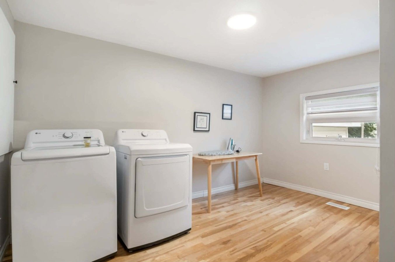 A laundry room with two white washing machines, a wooden table with an ironing board, and a window with blinds.