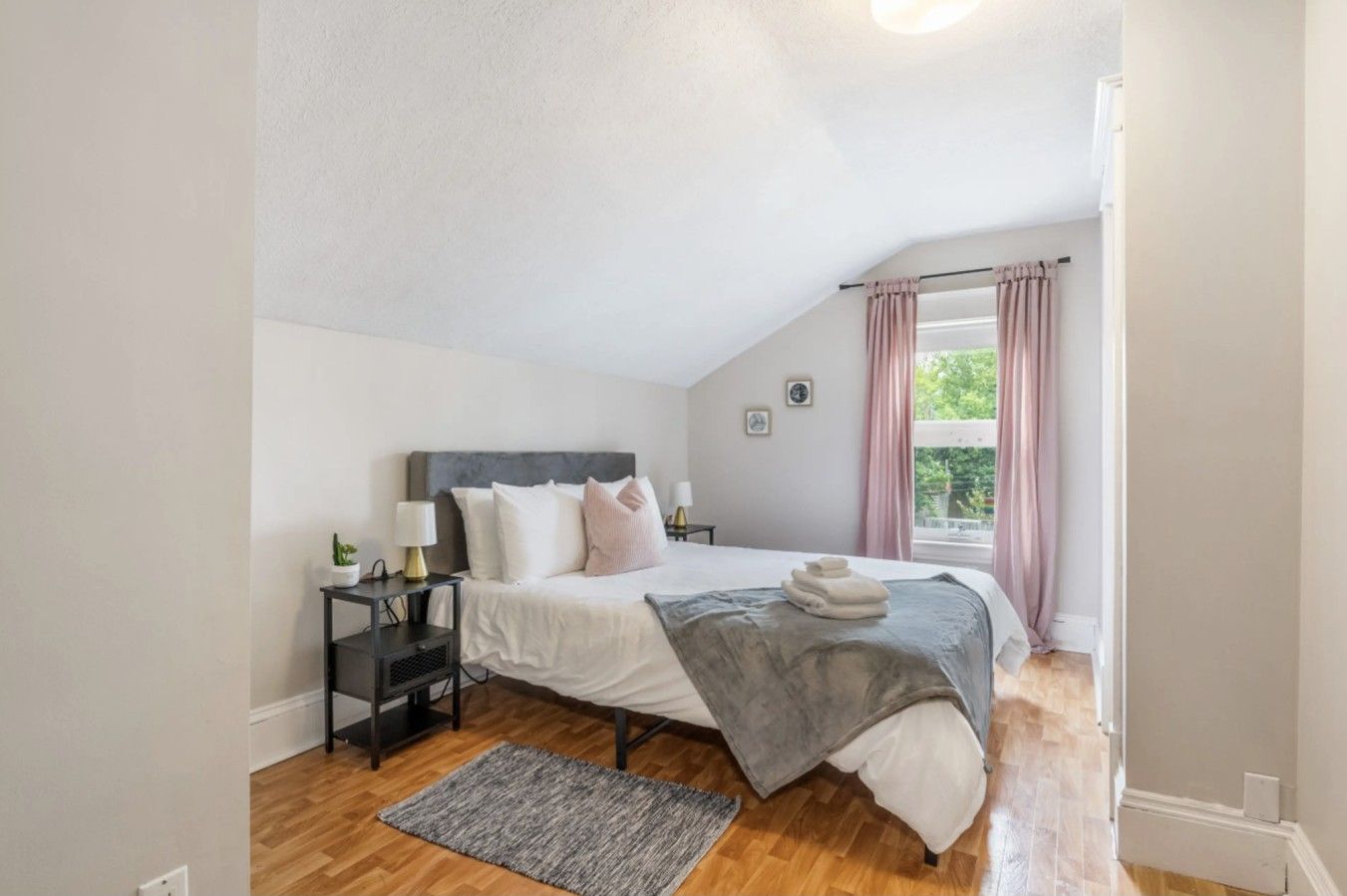 A bedroom with a bed, bedside tables, a nightstand, a window with curtains, and a view of greenery outside.