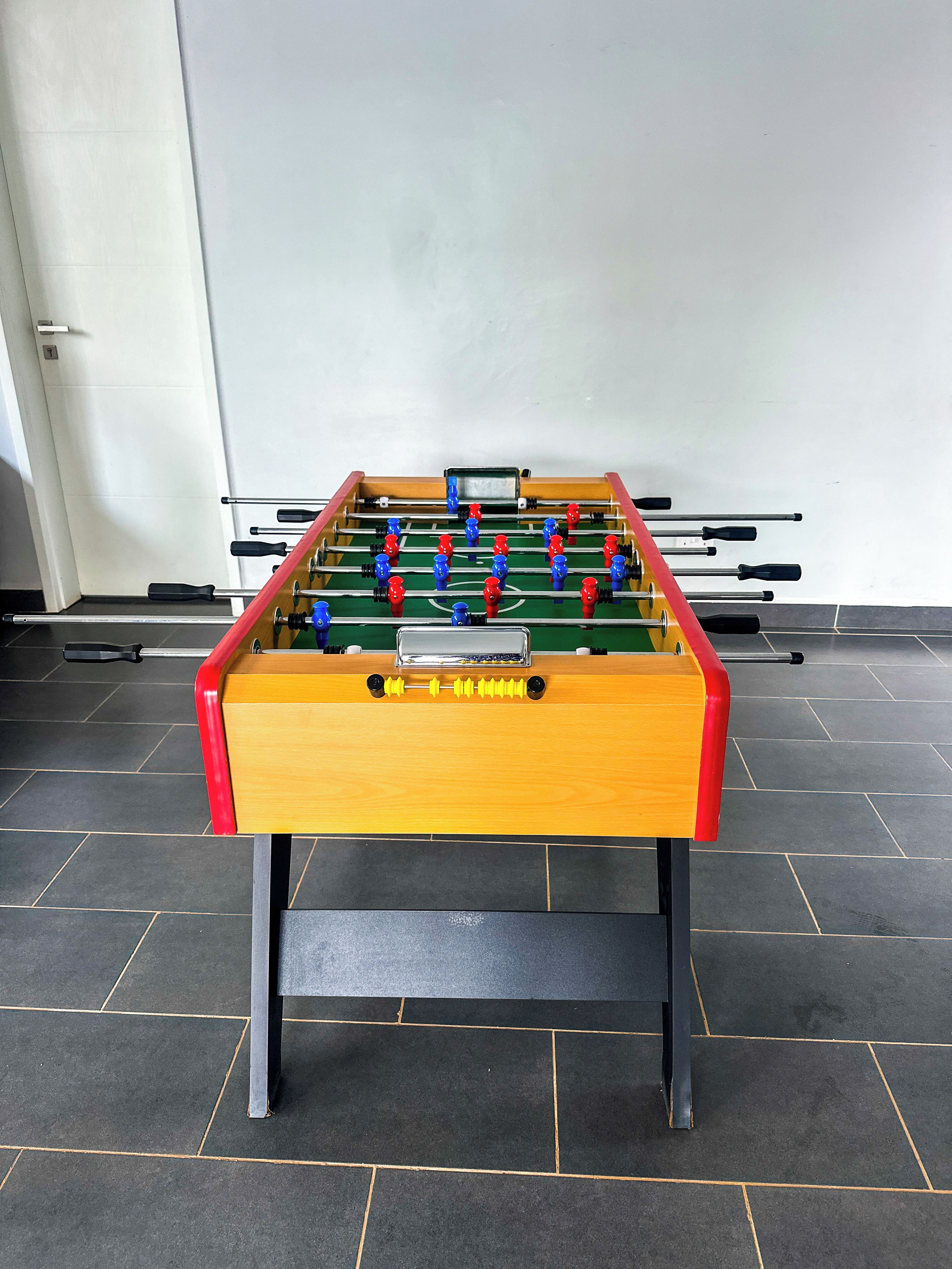 Une table de baby-foot avec un socle en bois et des côtés rouges et jaunes, posée sur un sol carrelé.