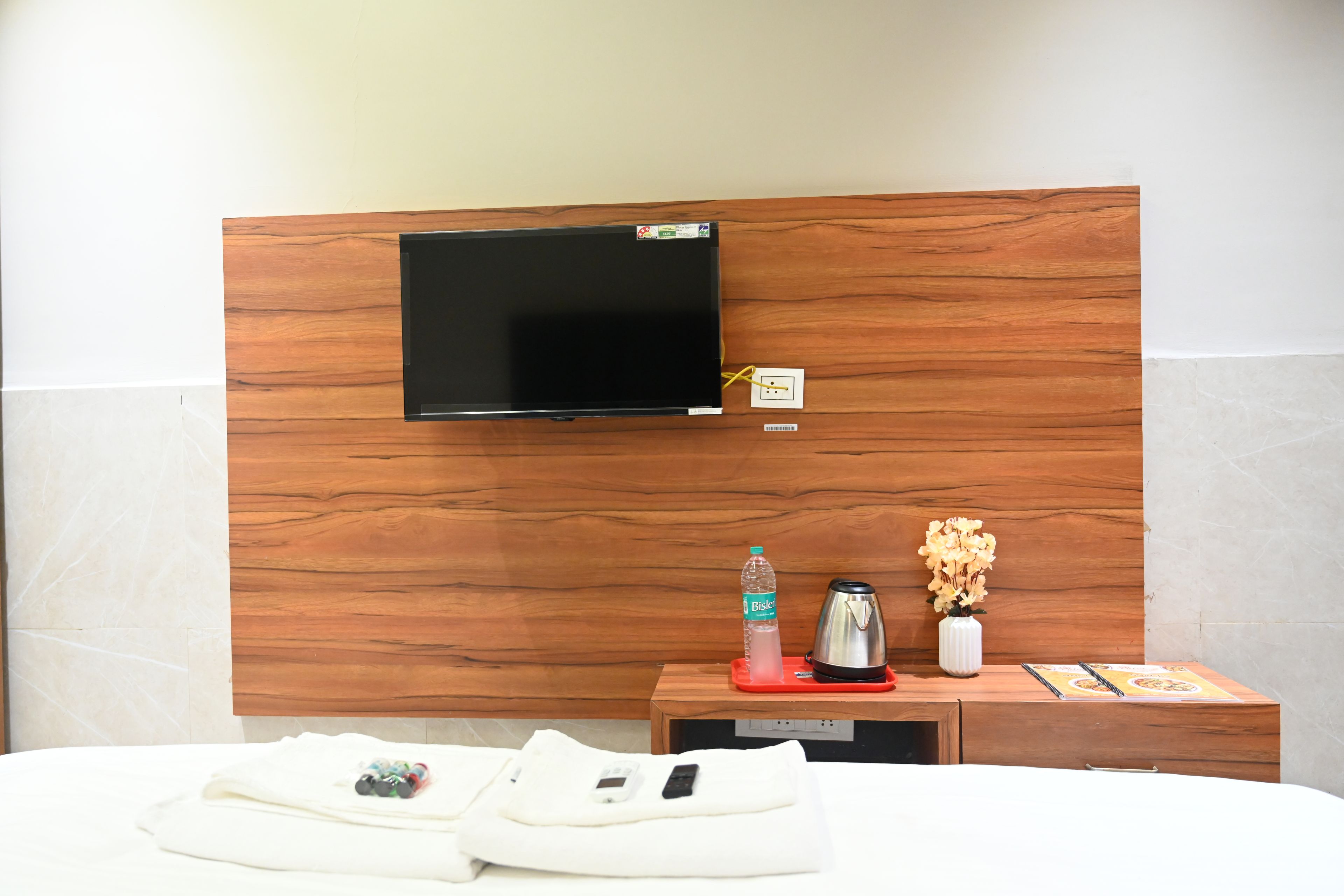 Une chambre d’hôtel avec une tête de lit en bois, un téléviseur à écran plat fixé au-dessus, une table de chevet avec une bouteille d’eau, une bouilloire et un vase de fleurs.