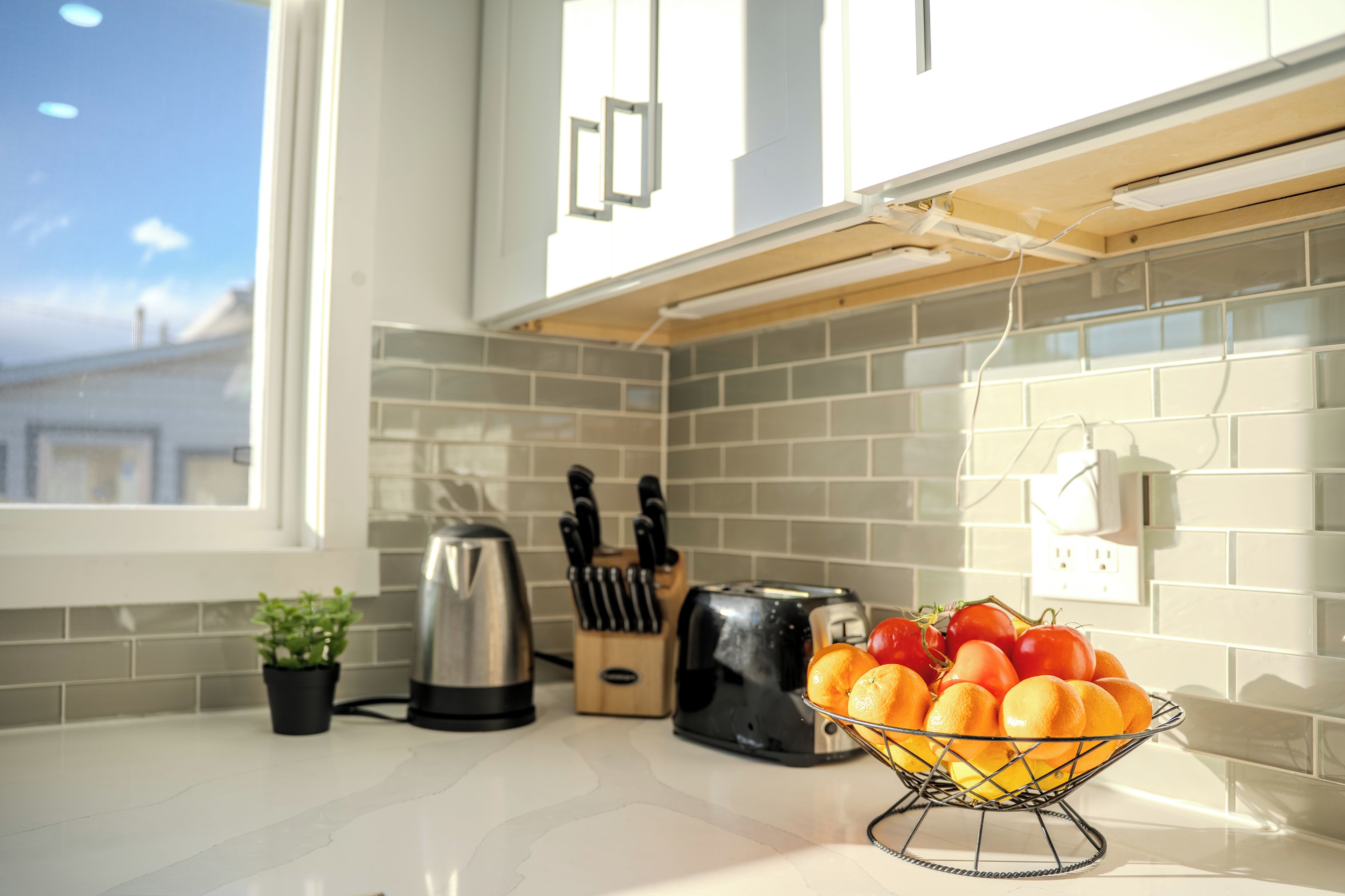 Une cuisine avec une fenêtre, un grille-pain, une bouilloire, un bloc à couteaux et un panier à fruits sur le plan de travail.