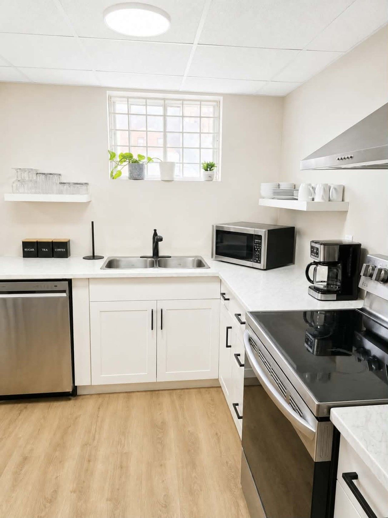 A modern kitchen with white cabinetry, a stainless steel dishwasher, a microwave, a coffee maker, and a window with plants.
