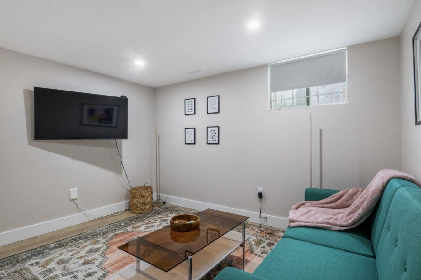 A living room with a sofa, a coffee table, a flat-screen TV mounted on the wall, and a window with blinds.