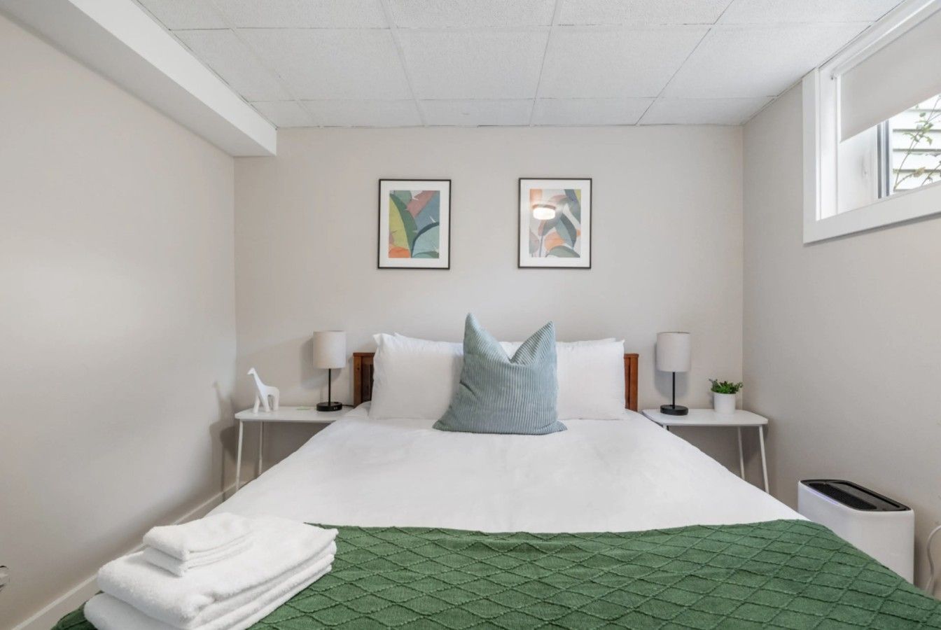 A hotel room with a bed, two bedside lamps, a green quilted bedspread, and two framed artworks on the wall.