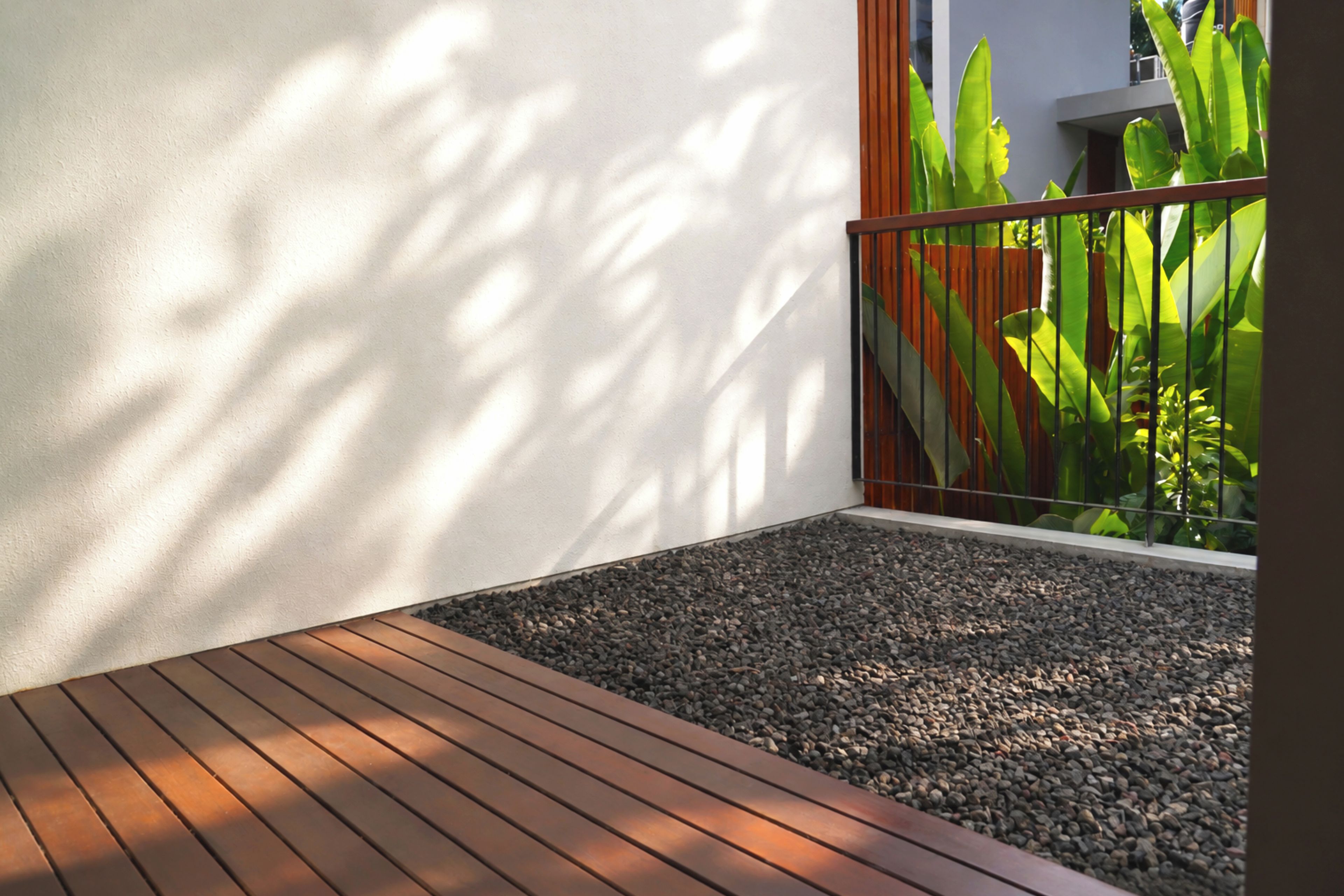A wooden deck with a gravel area, a white wall, and a metal railing with green foliage in the background.